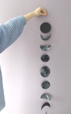 Painted moon phase wall art on a light gray wall with a person's arm in a blue sweater. A stitched paper garland as a living room wall hanging