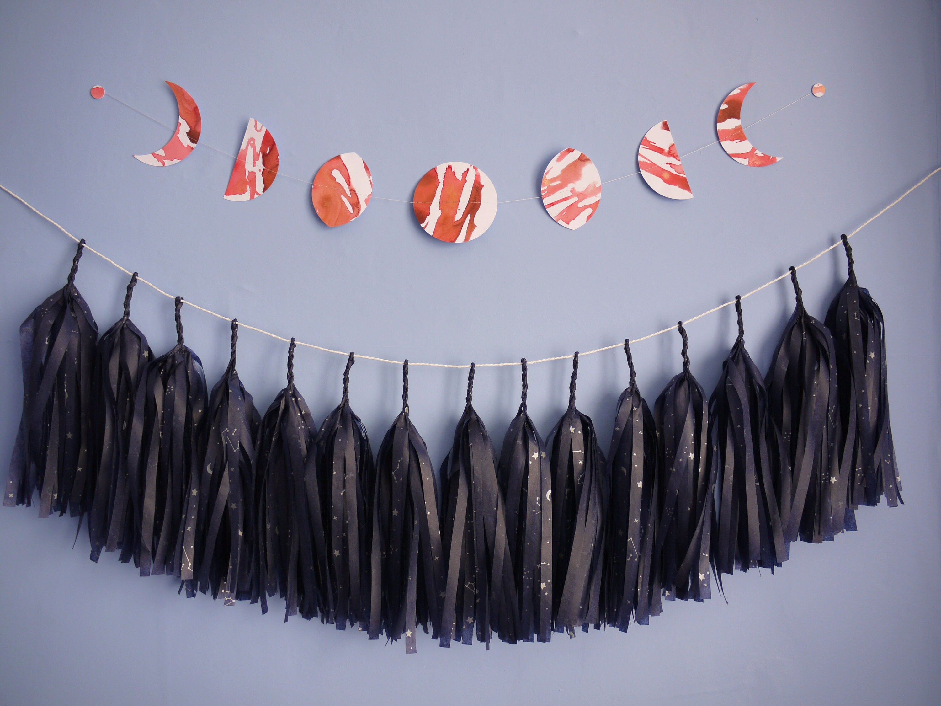 Red and white moon phases garland hung horizontally above navy tissue tassel garland with constellation pattern