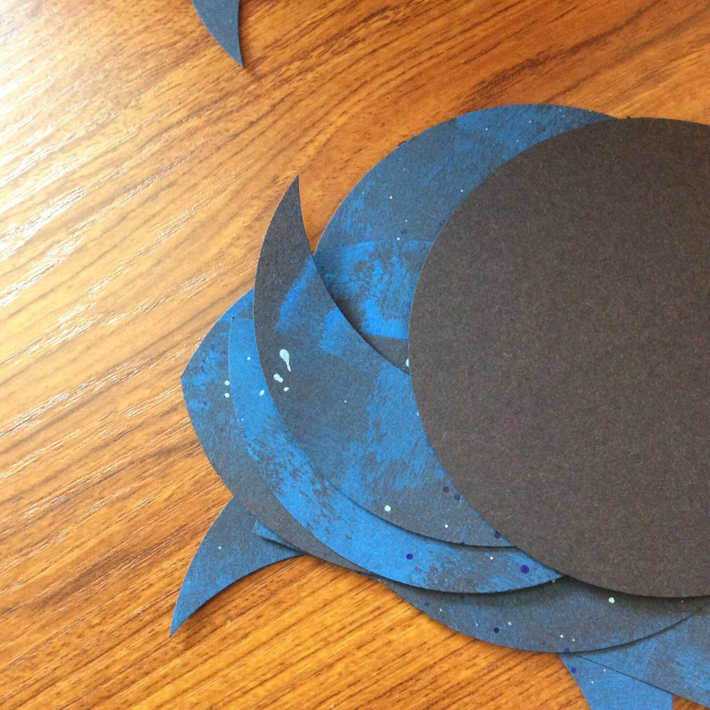 Blue and black circular paper pieces on a wooden surface. Moon phase shapes before becoming a stitched moon phases wall hanging