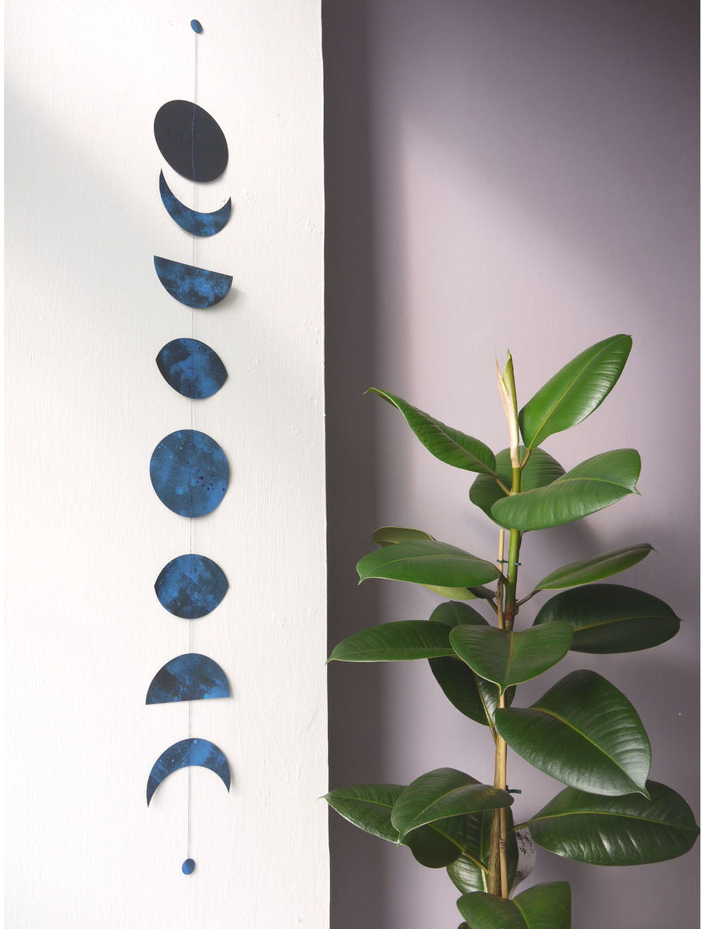 Decorative moon phase mobile hanging on a white wall with a plant in the foreground.
