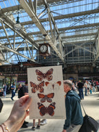 Person holding a risograph print with butterfly illustrations in busy Glasgow Central Station.
