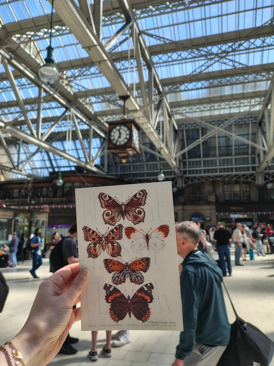 Person holding a risograph print with butterfly illustrations in busy Glasgow Central Station.