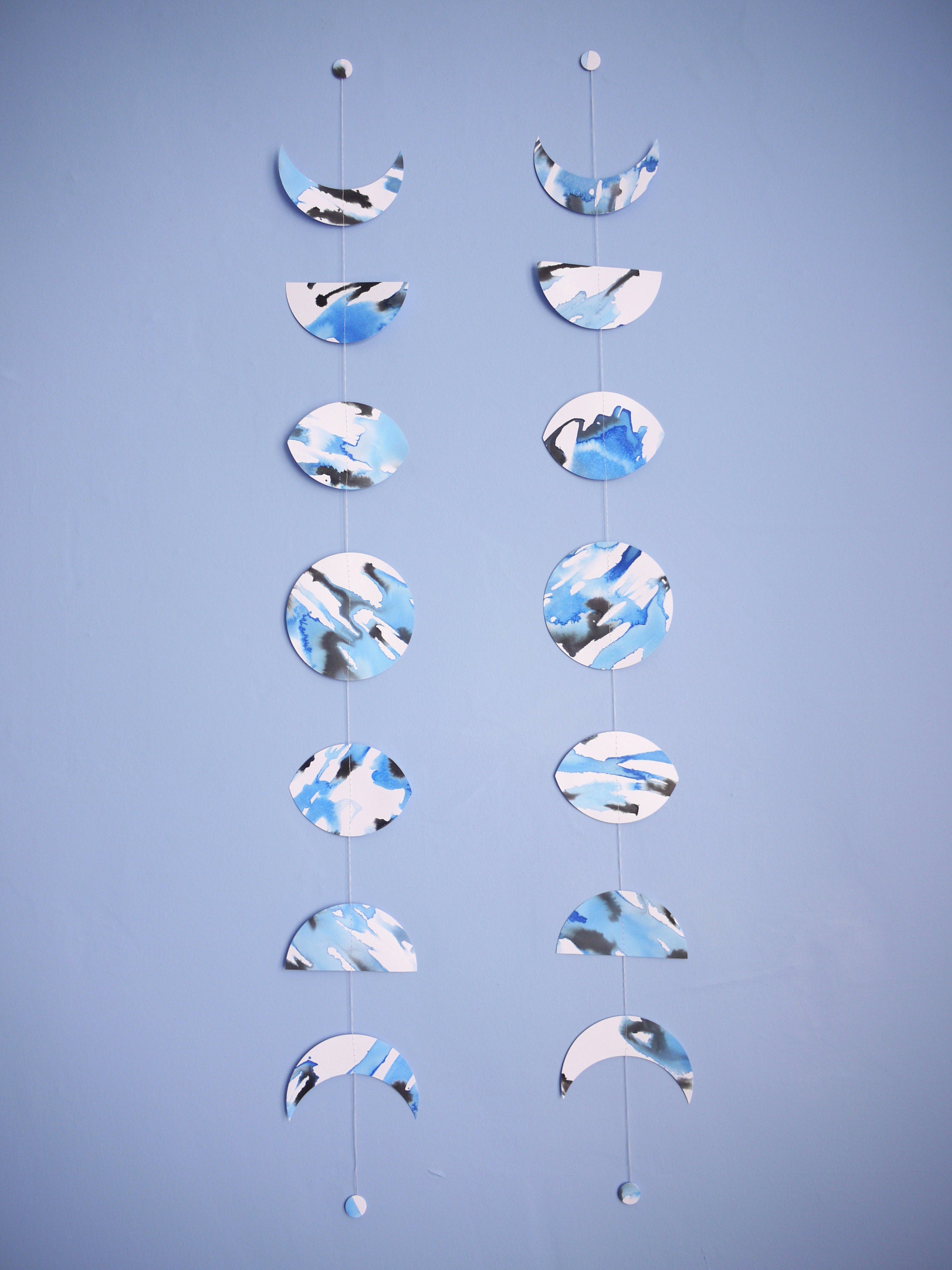 Two moon phase stitched garlands hanging vertically on a wall each painted slightly differently with blue and black ink displayed on a blue background 