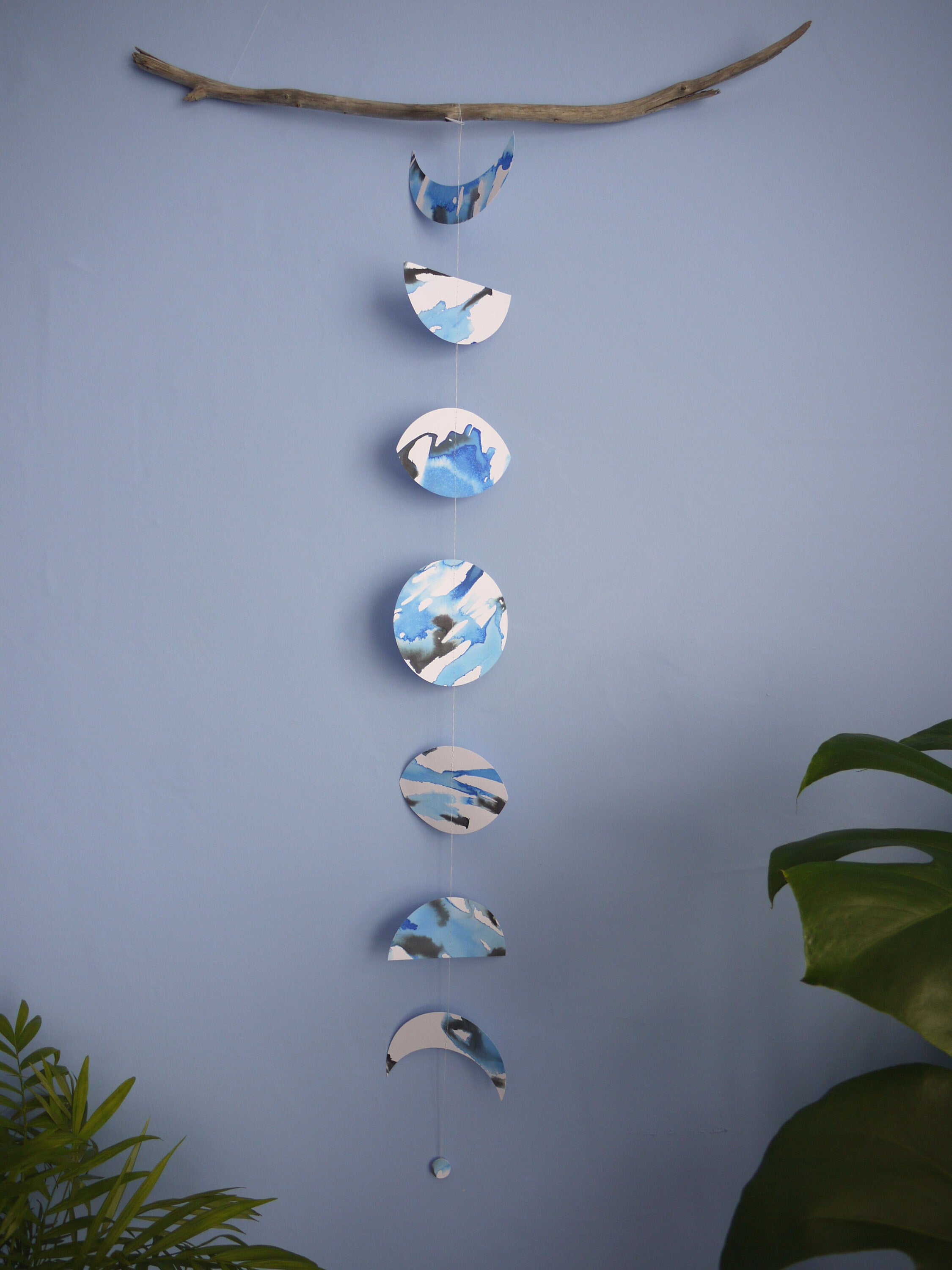 Paper garland of phases of the moon painted with blue and black ink stitched to create a wall hanging displayed on a blue background with plants either side