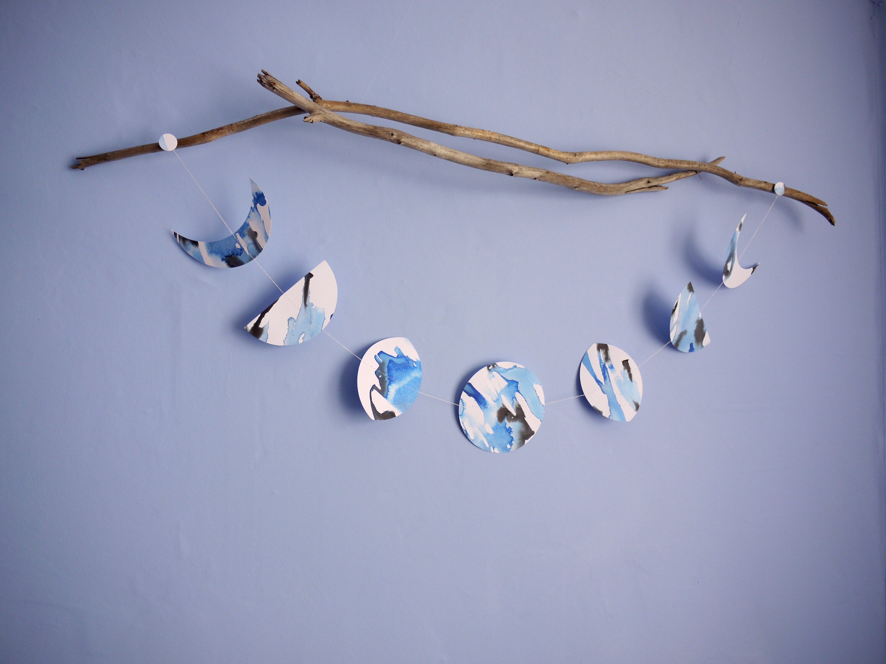 Moon phase shape stitched paper garland hung horizontally on driftwood painted with blue and black ink displayed on a blue background 