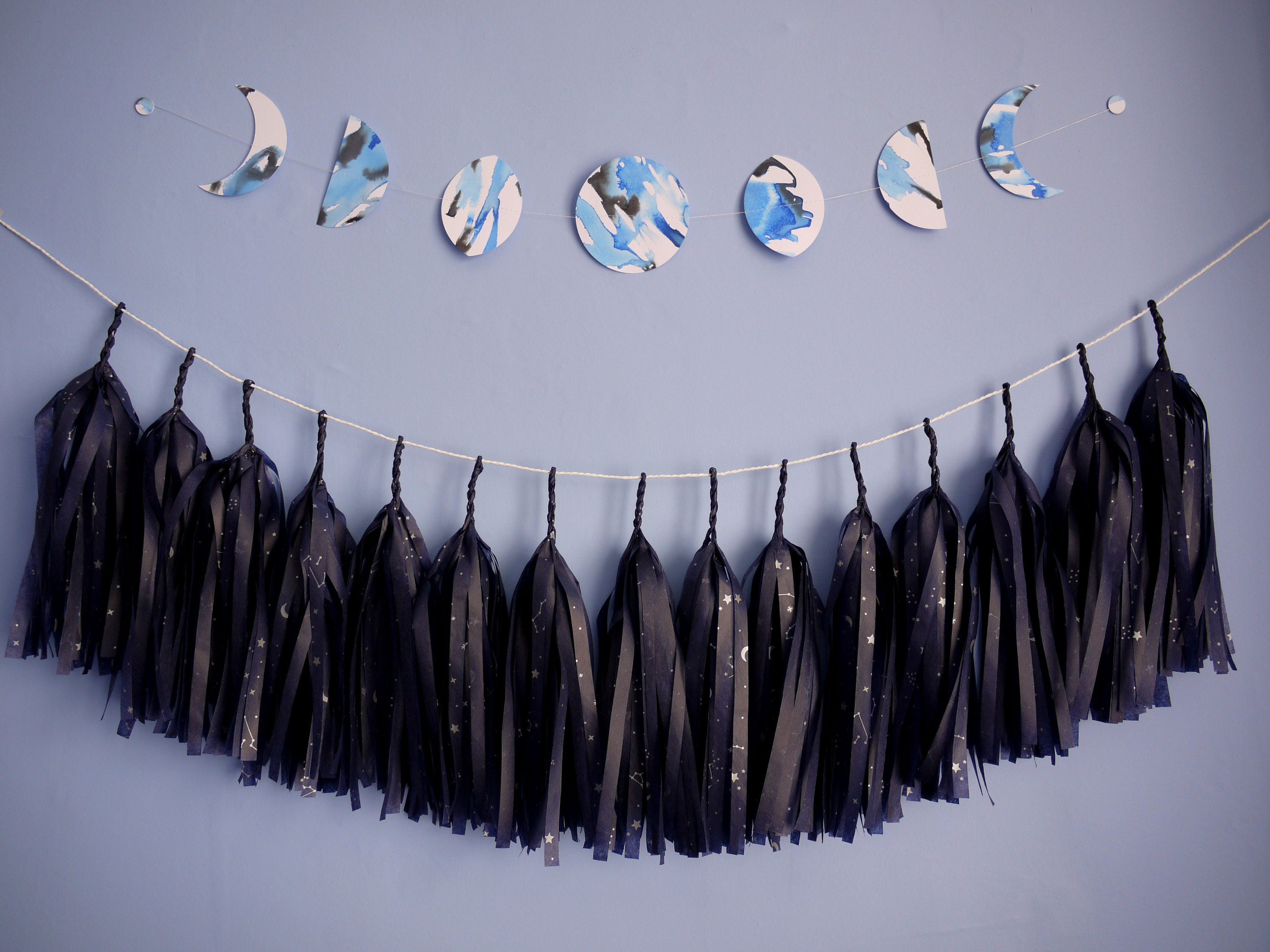 Blue and black ink painted on stitched moon phase shaped wall hanging painted with a strand of navy blue constellation printed tissue tassels hung underneath 