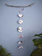 Vertical moon phases stitched paper garland painted with black blue and purple inks hanging  on a blue wall with green plants on either side