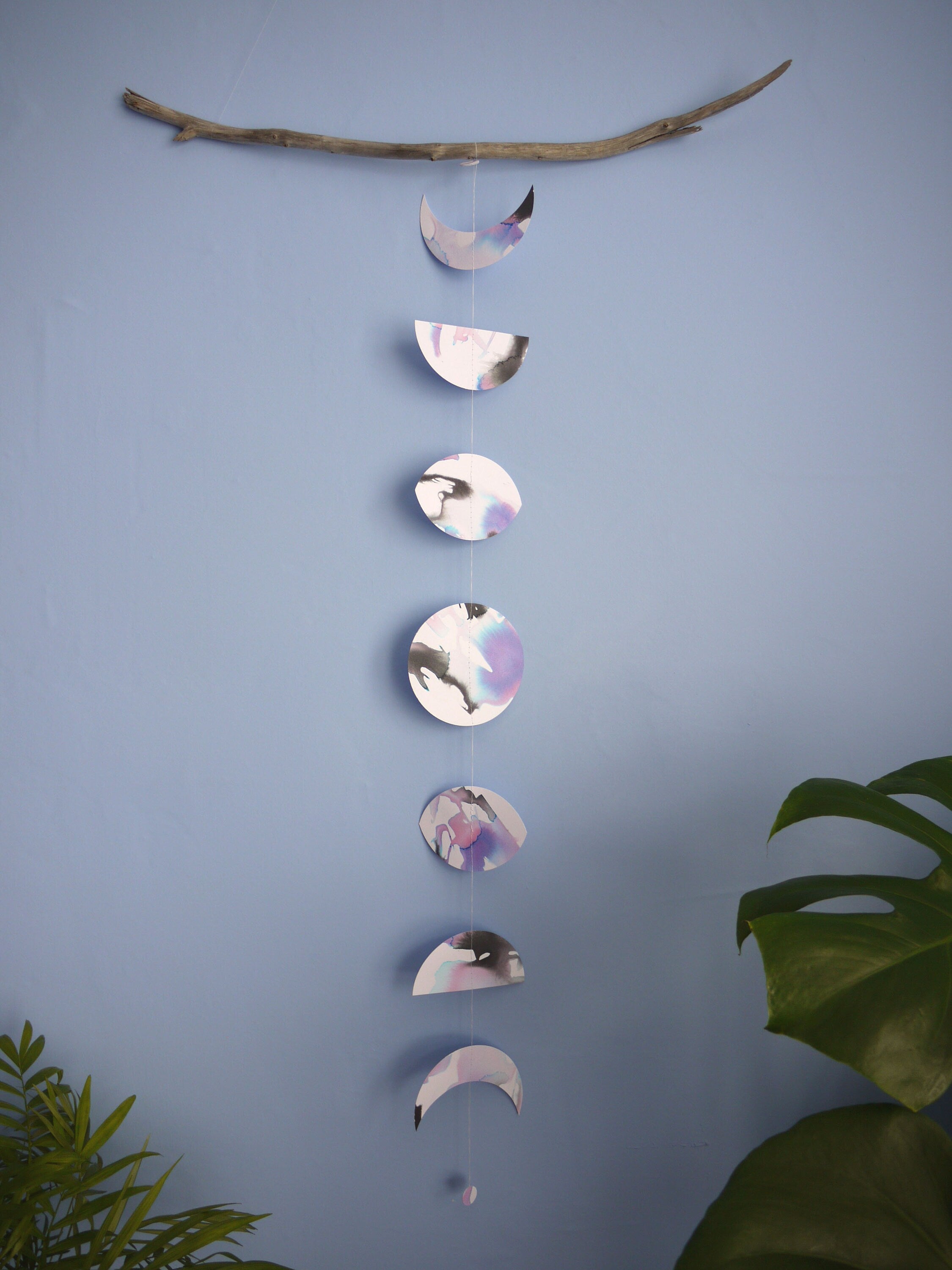 Vertical moon phases stitched paper garland painted with black blue and purple inks hanging  on a blue wall with green plants on either side