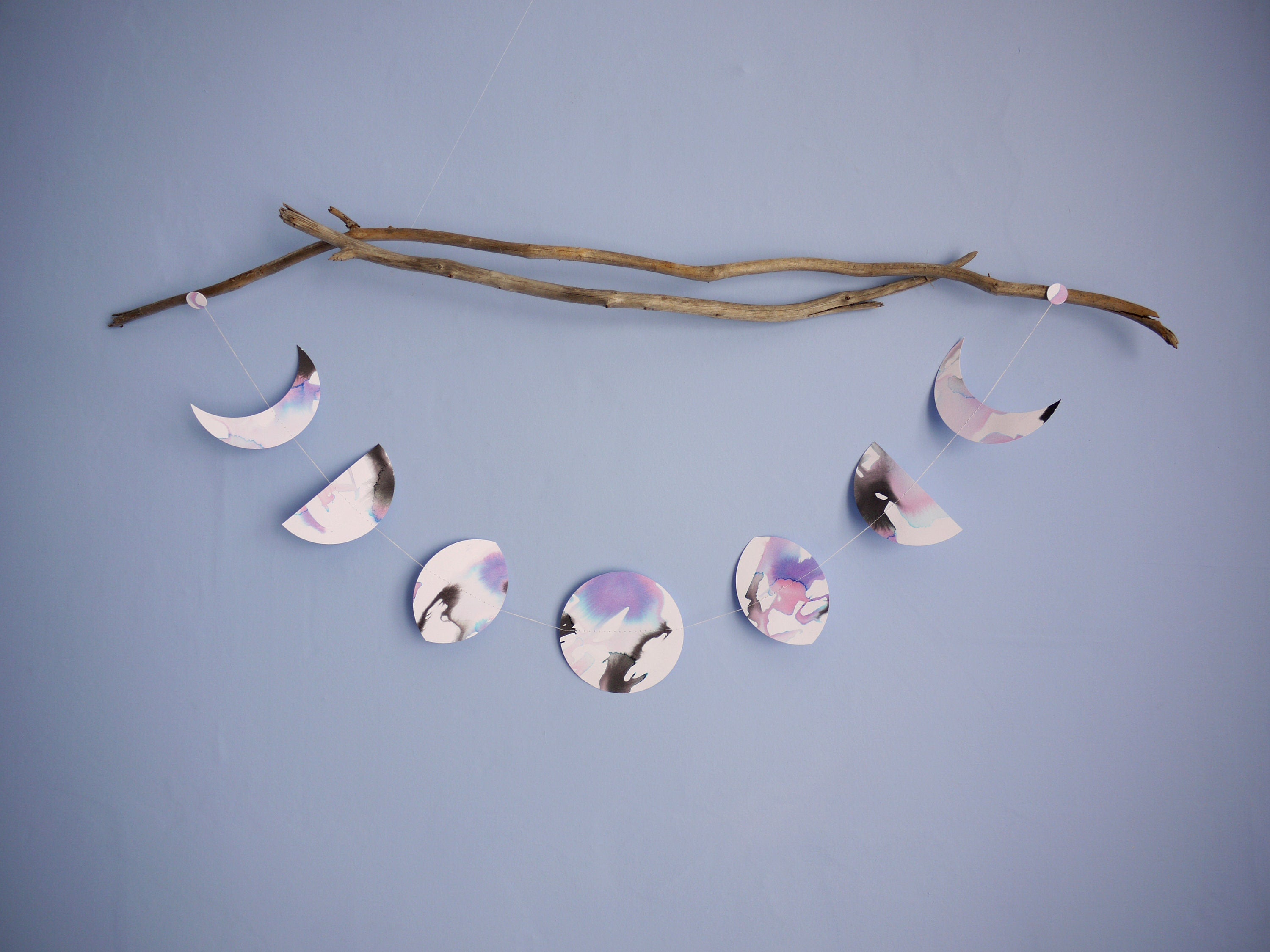 Horizontal moon phases shapes hanging as a stitched paper garland painted with black blue and purple inks from a piece of driftwood on a blue wall