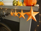 Fun easy halloween crafts. Whimsigoth orange and black paper star garland hanging across a gray fireplace mantel, with warm string lights and fall decorations like pumpkins and gourds in the background.