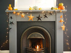 A gray fireplace mantel decorated with orange and black paper star garlands, warm white string lights, and small pumpkins, with a lit fire visible in the fireplace below. Whimsigoth fun easy halloween crafts.