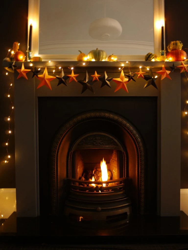 A cozy fireplace mantel decorated with warm white string lights, orange and black DIY paper star garland, small pumpkins, and candles, with a fire burning in the arched fireplace opening. Whimsigoth, fun easy halloween crafts.