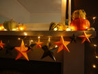 Fun easy halloween crafts with DIY paper star garland kit. Orange and black 3D paper stars with warm white string lights, displayed on a mantelpiece alongside pumpkins and gourds.