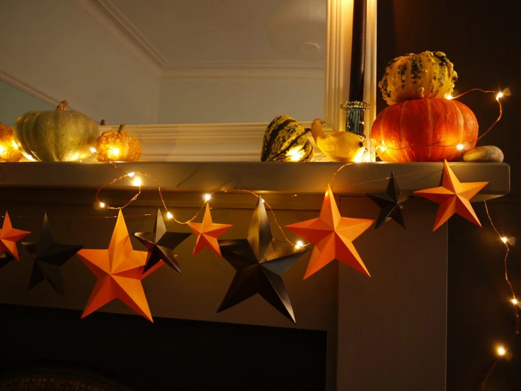 Fun easy halloween crafts with DIY paper star garland kit. Orange and black 3D paper stars with warm white string lights, displayed on a mantelpiece alongside pumpkins and gourds.