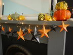 A whimsigoth Halloween mantel display featuring orange and black 3D DIY paper star garland, warm string lights, pumpkins, and gourds against a light blue wall. Fun easy halloween crafts.