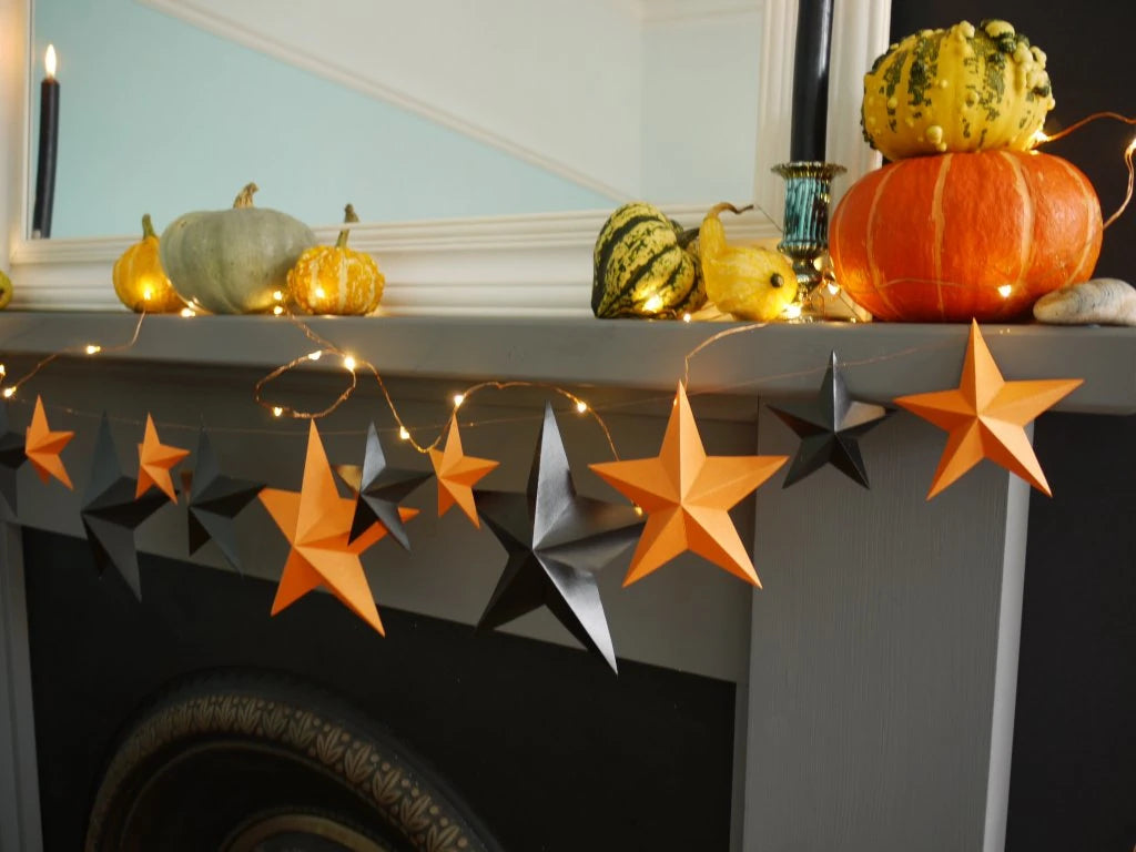 A whimsigoth Halloween mantel display featuring orange and black 3D DIY paper star garland, warm string lights, pumpkins, and gourds against a light blue wall. Fun easy halloween crafts.