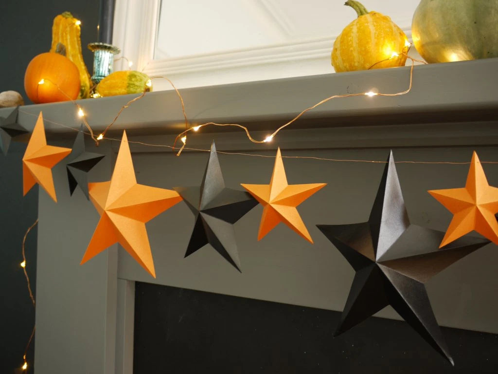 Orange and black 3D paper star garland hanging across a gray fireplace mantel, styled with warm string lights and autumn pumpkins.