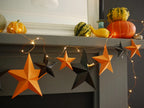Orange and black 3D DIY star paper garland with warm string lights draped over a gray mantel, styled with pumpkins and gourds in a home setting. Fun easy Halloween crafts for whimsigoth home decor.