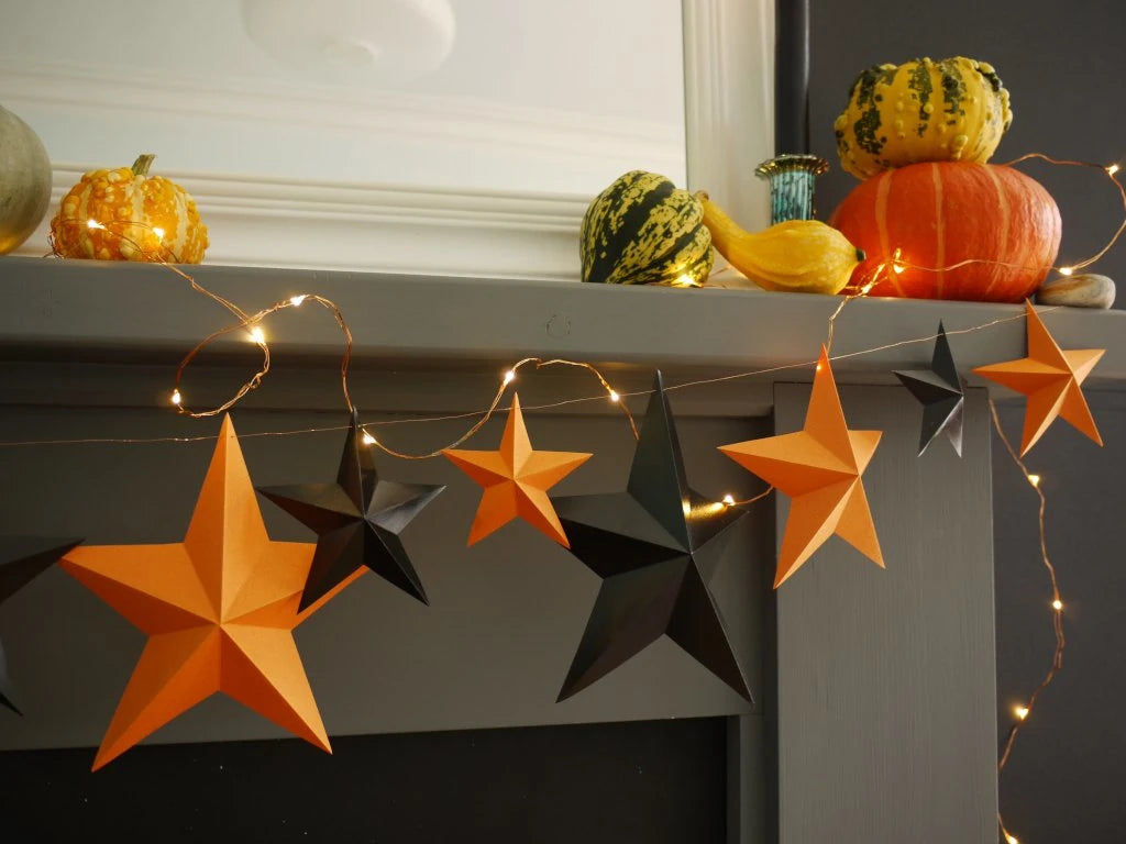 Orange and black 3D DIY star paper garland with warm string lights draped over a gray mantel, styled with pumpkins and gourds in a home setting. Fun easy Halloween crafts for whimsigoth home decor.
