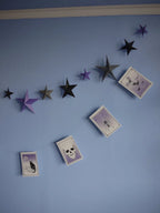Halloween-themed wall decor featuring a DIY paper star garland of 3D paper stars in black, gray, and purple, alongside a riso print paper garland with spooky illustrations like a cat, spider, bats and skull, displayed on a light blue wall. DIY Samhain decorations.