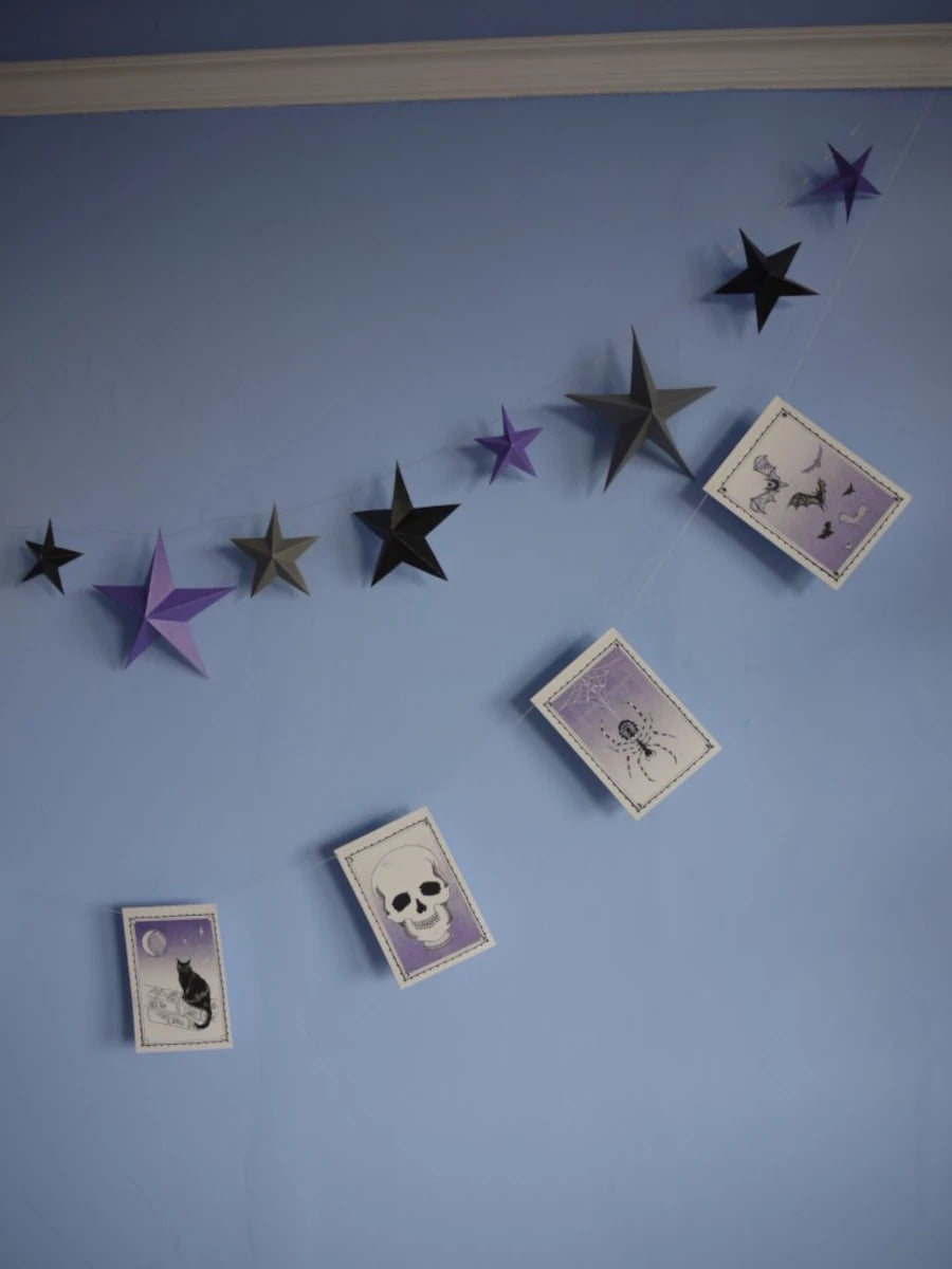 Halloween-themed wall decor featuring a DIY paper star garland of 3D paper stars in black, gray, and purple, alongside a riso print paper garland with spooky illustrations like a cat, spider, bats and skull, displayed on a light blue wall. DIY Samhain decorations.