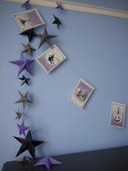Whimsigoth DIY Samhain decorations featuring a DIY paper star garland in black, gray, and purple, alongside four small square cards with spooky illustrations, displayed on a light blue wall.