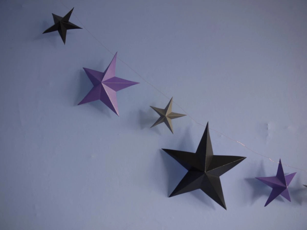 Whimsigoth DIY Samhain decorations. DIY paper star garland of 3D stars in black, purple, and pewter paper hanging against a light blue wall.