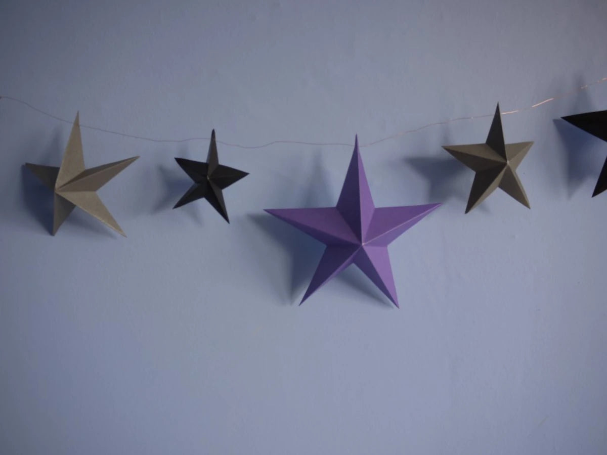 Whimsigoth DIY Samhain decorations. DIY paper star garland of 3D stars in black, purple, and pewter paper hanging against a light blue wall.