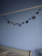 Whimsigoth DIY Samhain decorations. A DIY paper star garland of 3D stars in black, purple, and pewter paper hanging across a light blue bedroom wall above a bed.