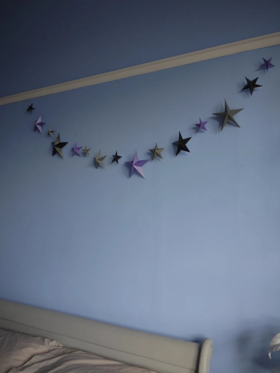 Whimsigoth DIY Samhain decorations. A DIY paper star garland of 3D stars in black, purple, and pewter paper hanging across a light blue bedroom wall above a bed.