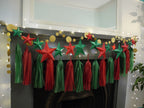 DIY paper star making for Christmas. A festive Christmas garland with red and green paper stars and matching hanging tassels, draped across a fireplace mantel with gold circular garland and warm string lights.