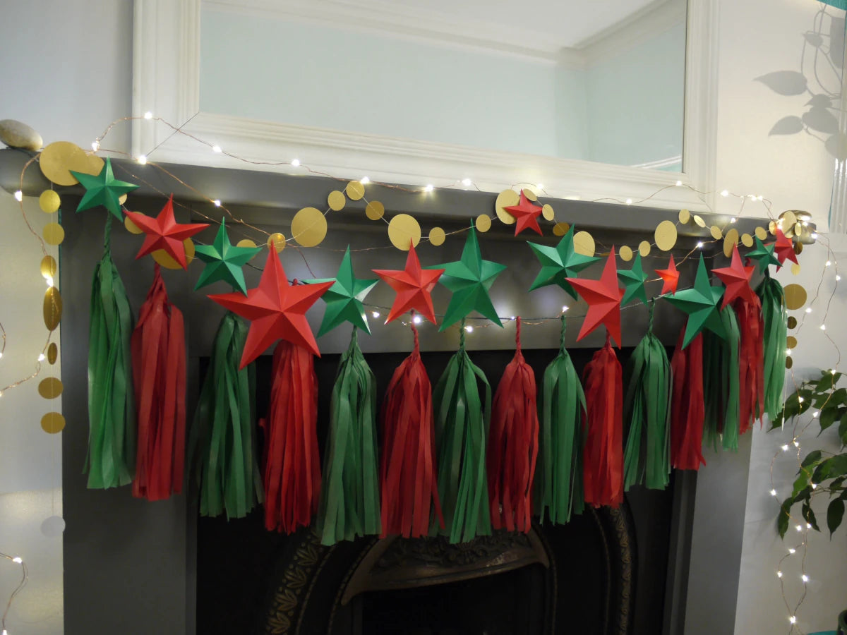 DIY paper star making for Christmas. A festive Christmas garland with red and green paper stars and matching hanging tassels, draped across a fireplace mantel with gold circular garland and warm string lights.