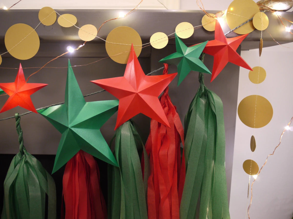 DIY paper star making for Christmas. Christmas holiday garland decoration featuring red and green paper star garland and matching red and green paper tassels, with gold circular stitched paper garland and warm string lights.