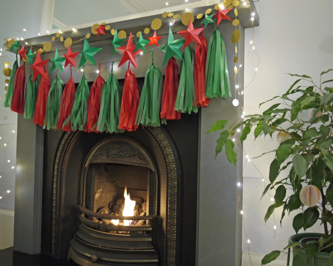 DIY paper star making for Christmas. A festive Christmas garland with red and green paper tassels and a DIY paper star garland hanging above a fireplace with a fire burning inside. White string lights are draped around the fireplace and a green potted plant is visible on the right.
