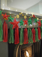 DIY paper star making for Christmas. A festive Christmas garland hanging above a fireplace, featuring red and green paper star garland, matching paper tassels, and gold circular sewn paper garland with warm string lights.