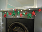 DIY paper star making for Christmas. A string of red and green paper star garlands with warm white fairy lights draped across a dark fireplace mantel in an indoor room.