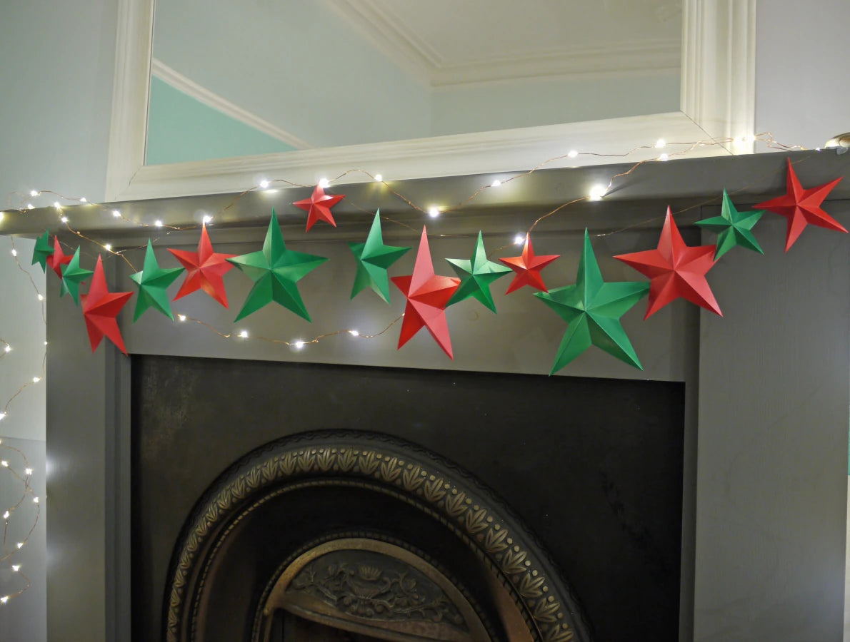 DIY paper star making for Christmas. A string of red and green paper star garlands with warm white fairy lights draped across a dark fireplace mantel in an indoor room.
