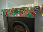 DIY paper star making for Christmas. A festive Christmas garland with red and green paper star garland and gold circular sewn paper garland, strung with warm white fairy lights above a dark fireplace mantel.