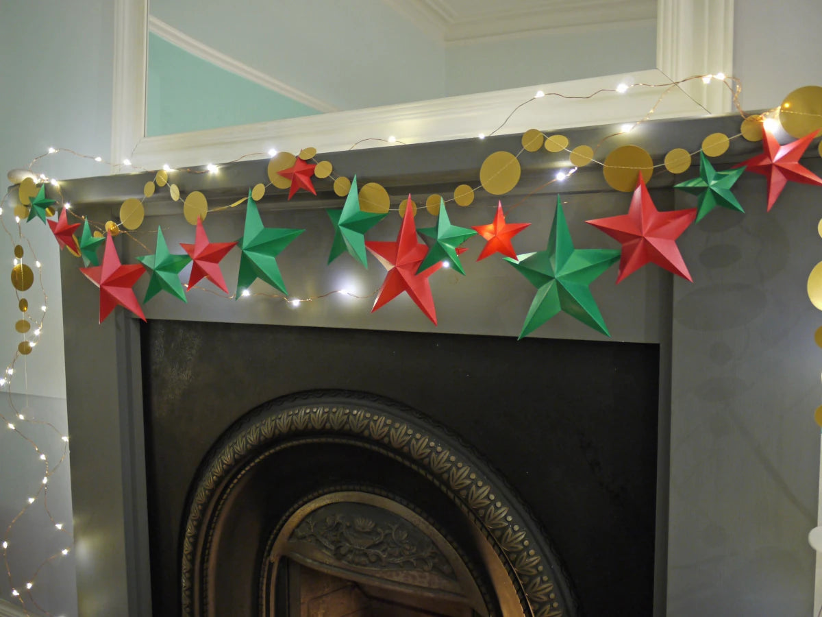 DIY paper star making for Christmas. A festive Christmas garland with red and green paper star garland and gold circular sewn paper garland, strung with warm white fairy lights above a dark fireplace mantel.