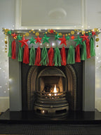 DIY paper star making for Christmas. Christmas fireplace decoration featuring red and green paper tassels, red and green DIY paper star garland, and gold circular stitched paper garland draped across a gray fireplace mantel with a lit fire inside.