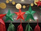 DIY paper star making for Christmas. Red and green paper star and tassel garland decorations hanging against a dark wall with warm string lights and gold circular garland above.
