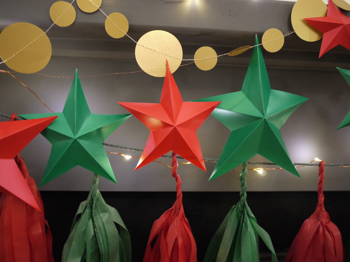 DIY paper star making for Christmas. Red and green paper star and tassel garland decorations hanging against a dark wall with warm string lights and gold circular garland above.