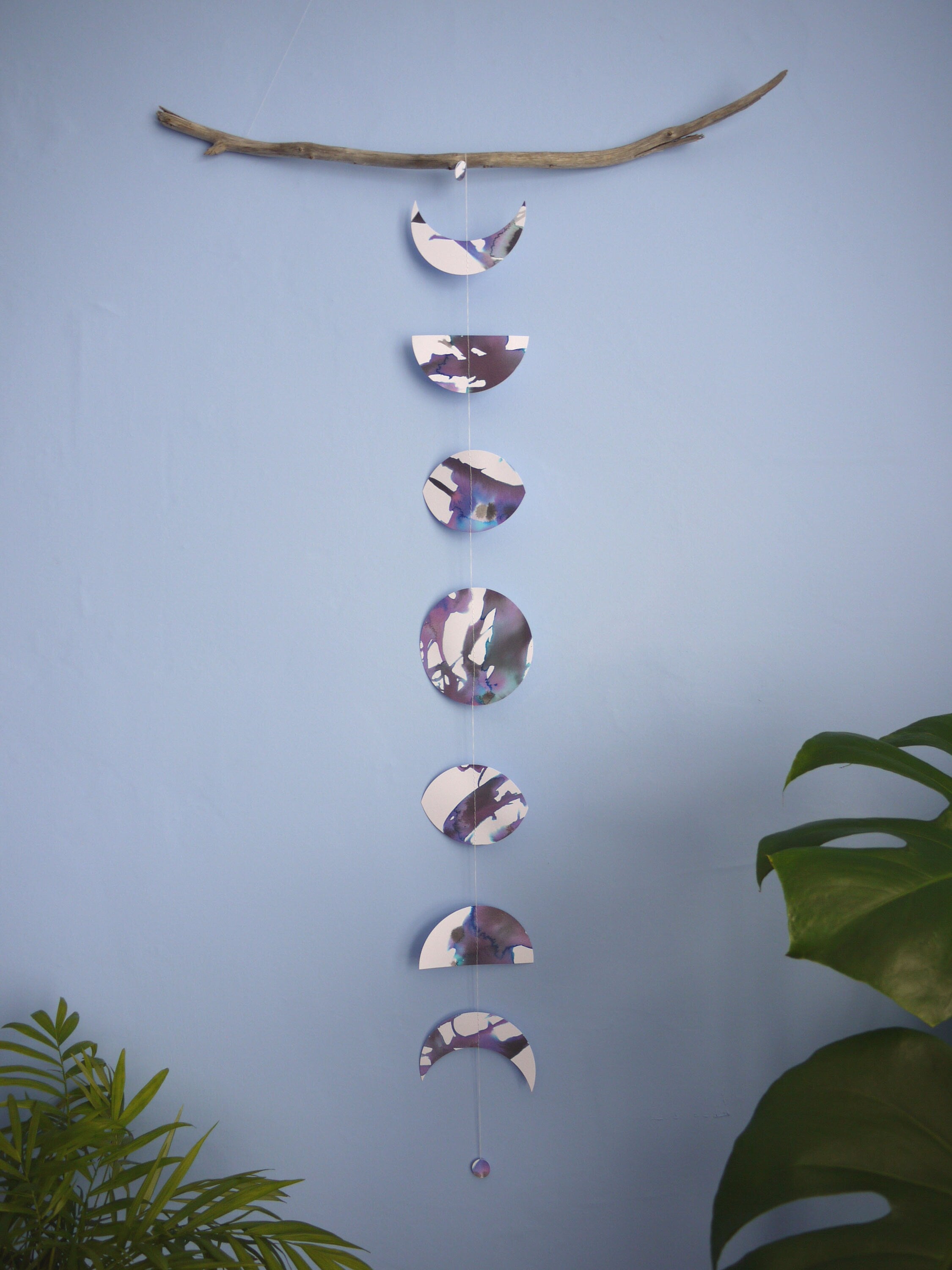 A purple, black and white moon phases wall hanging hanging vertically from driftwood, plants in the foreground