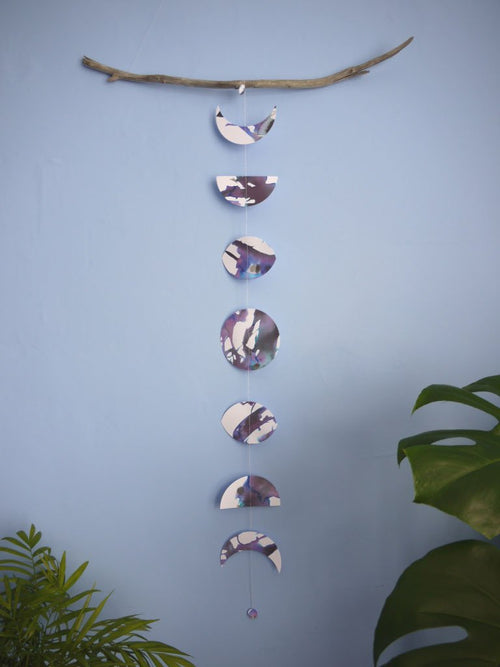 A purple, black and white moon phases wall hanging hanging vertically from driftwood, plants in the foreground