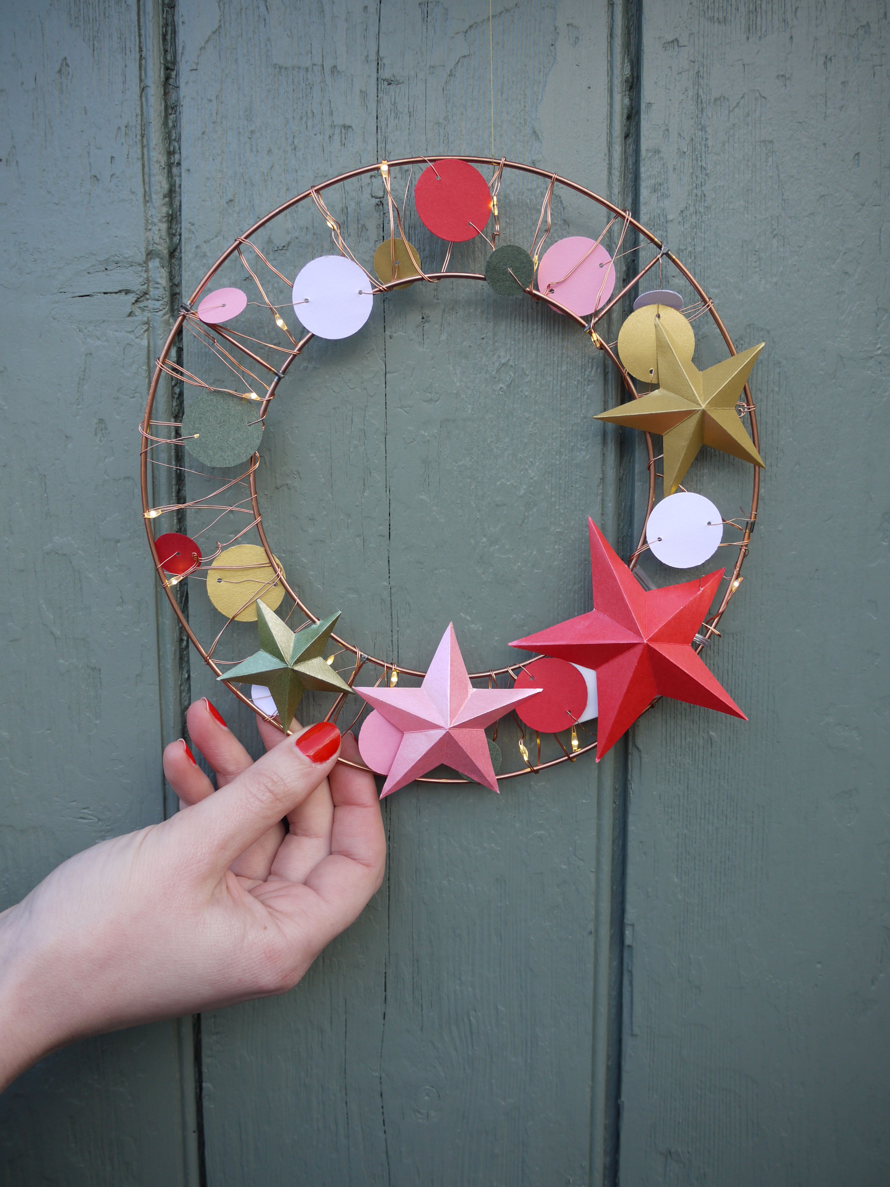Decorative wreath with colorful stars held by a hand against a wooden door.