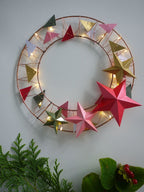 Decorative wreath with paper stars and string lights on a white wall.