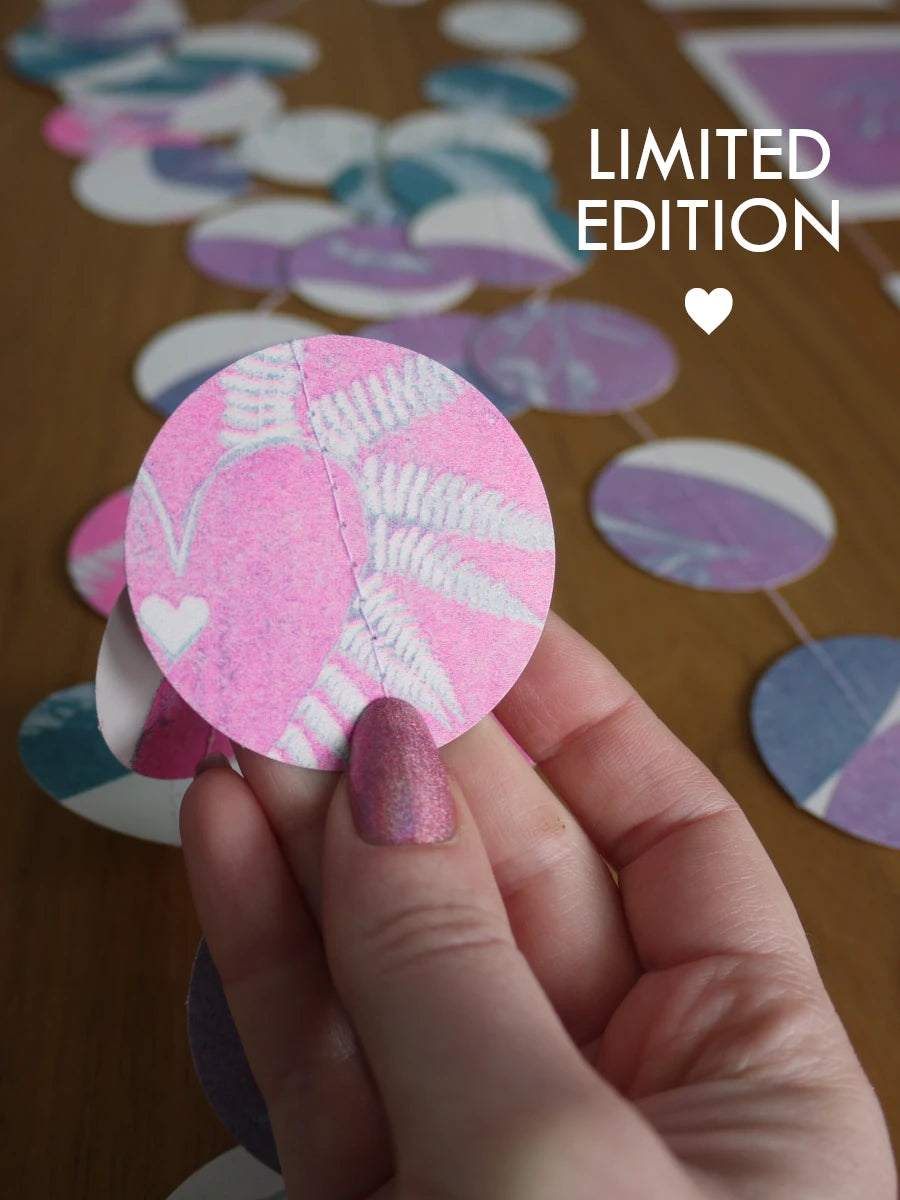 A hand with glittery pink nails holds a circular pink and white fern-patterned circle, with with the rest of the riso print garland scattered on a wooden surface in the background. White text in the upper right reads 'LIMITED EDITION' with a small heart.