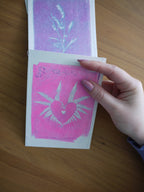 Everyday Enchantment - A hand with pink nail polish holds a small cyanotype riso print design at the bottome of a riso print wall hanging. The print features a white heart and fern design on a bright pink background, with another card showing a white plant on a purple background, placed on a wooden table.