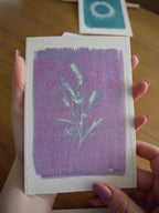 Everyday Enchantment - A person holds a small cyanotype riso print featuring a lavender-purple image. Another similar print is visible in the blurred background on a wooden surface.