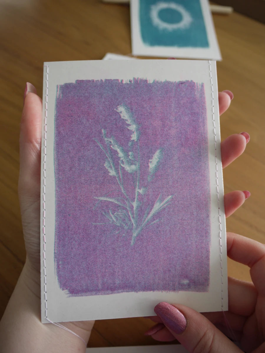 Everyday Enchantment - A person holds a small cyanotype riso print featuring a lavender-purple image. Another similar print is visible in the blurred background on a wooden surface.